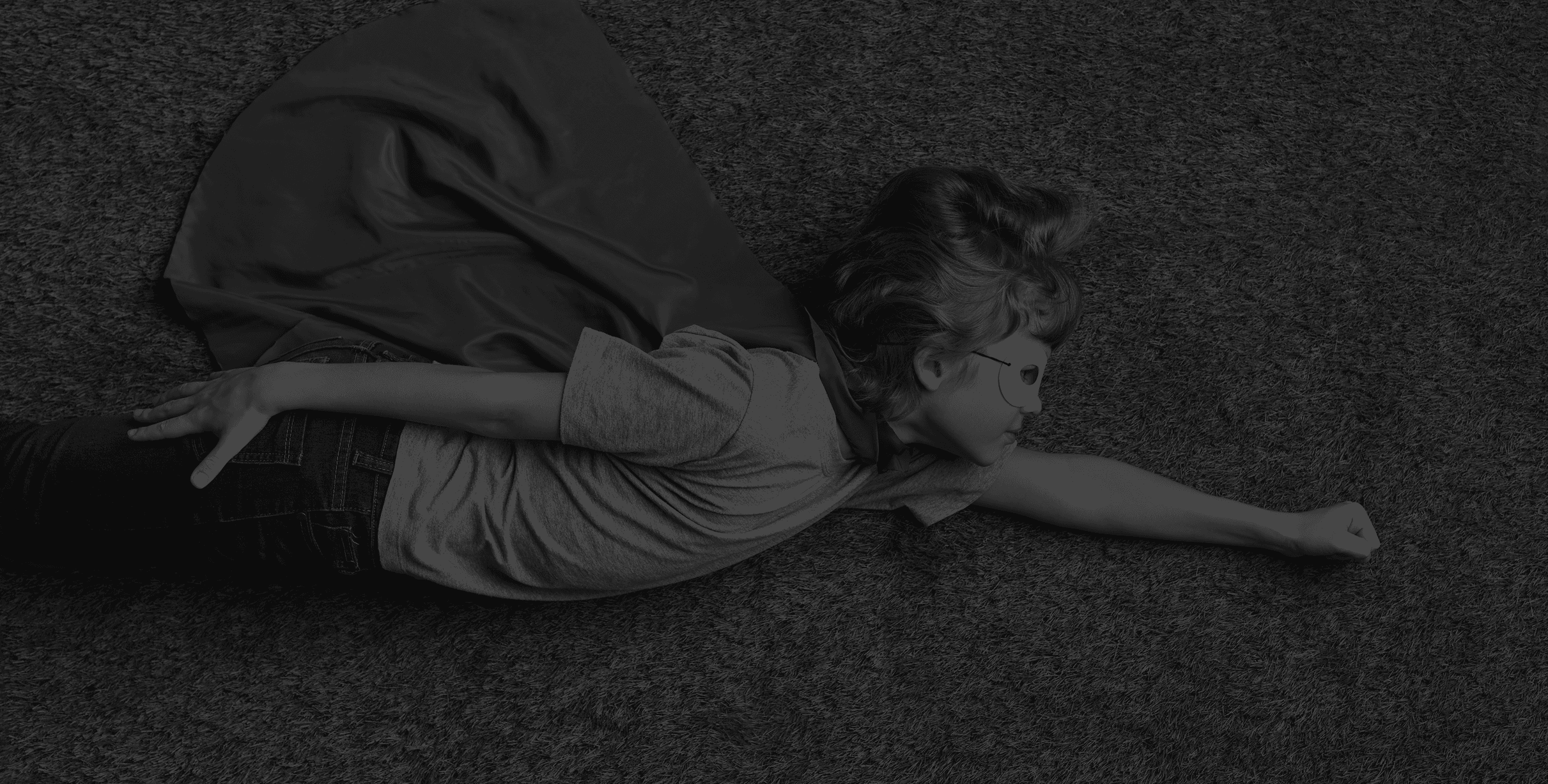 Black-and-white photo of a child in a cape lying on the floor with arms outstretched