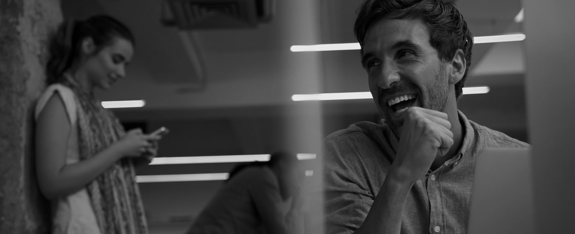 Black-and-white office photo of a laughing man with a colleague on her phone in the background